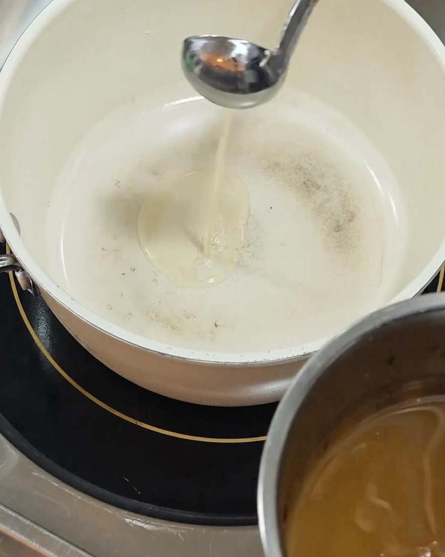 Un cucharón plateado vierte aceite amarillo claro en una olla blanca con revestimiento cerámico colocada sobre una placa de inducción negra.