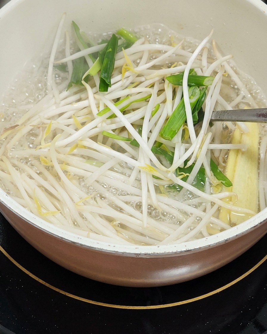 Una olla de agua hirviendo que contiene brotes de soja, hojas de ajo picadas y trozos de mazorquitas de maíz que se remueven con un utensilio de metal.