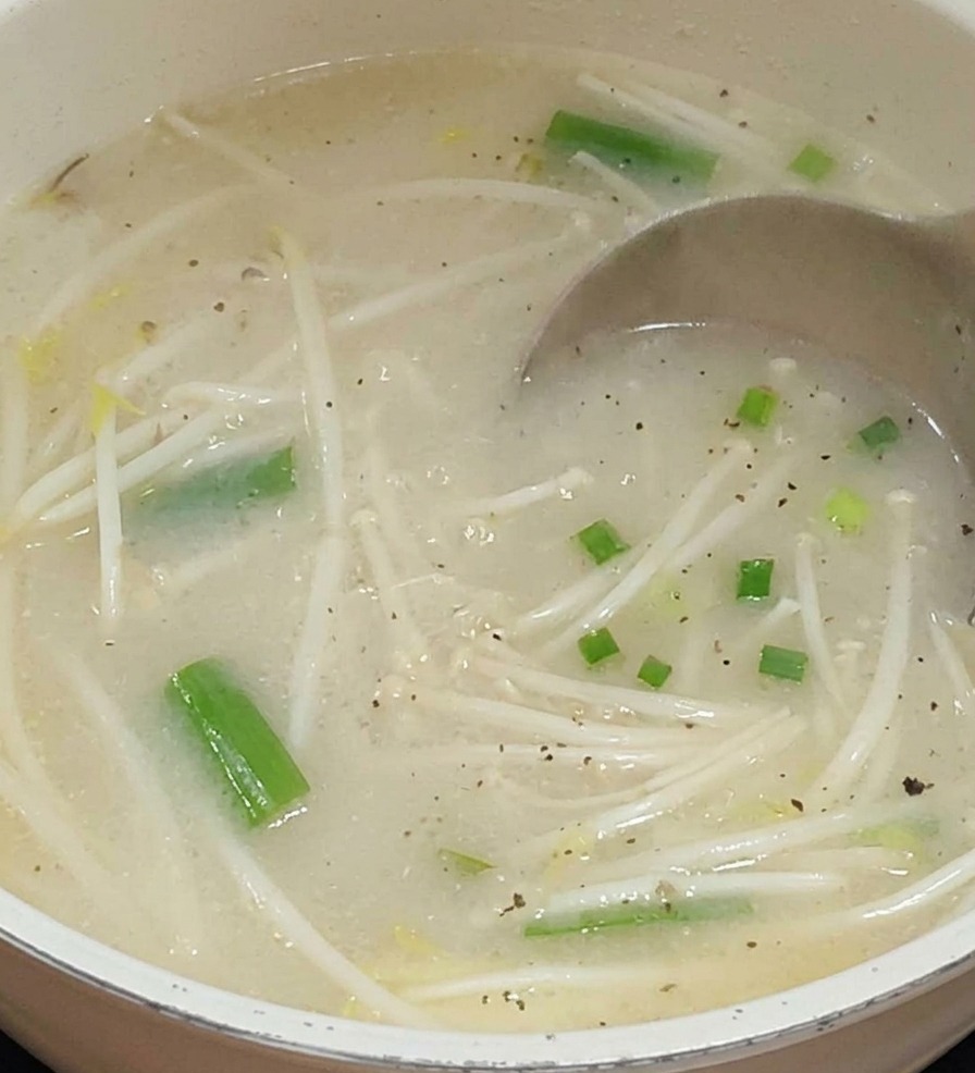 Un cucharón de metal vierte un caldo blanquecino y lechoso en una olla que contiene brotes de soja y verduras verdes.
