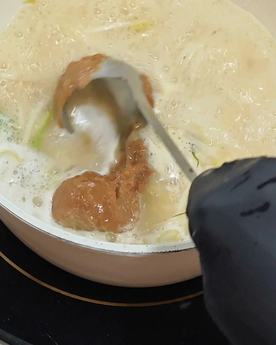 Una mano con guante usa un cucharón para remover una porción espesa de pasta de miso de color marrón rojizo en una base de sopa cremosa que burbujea.