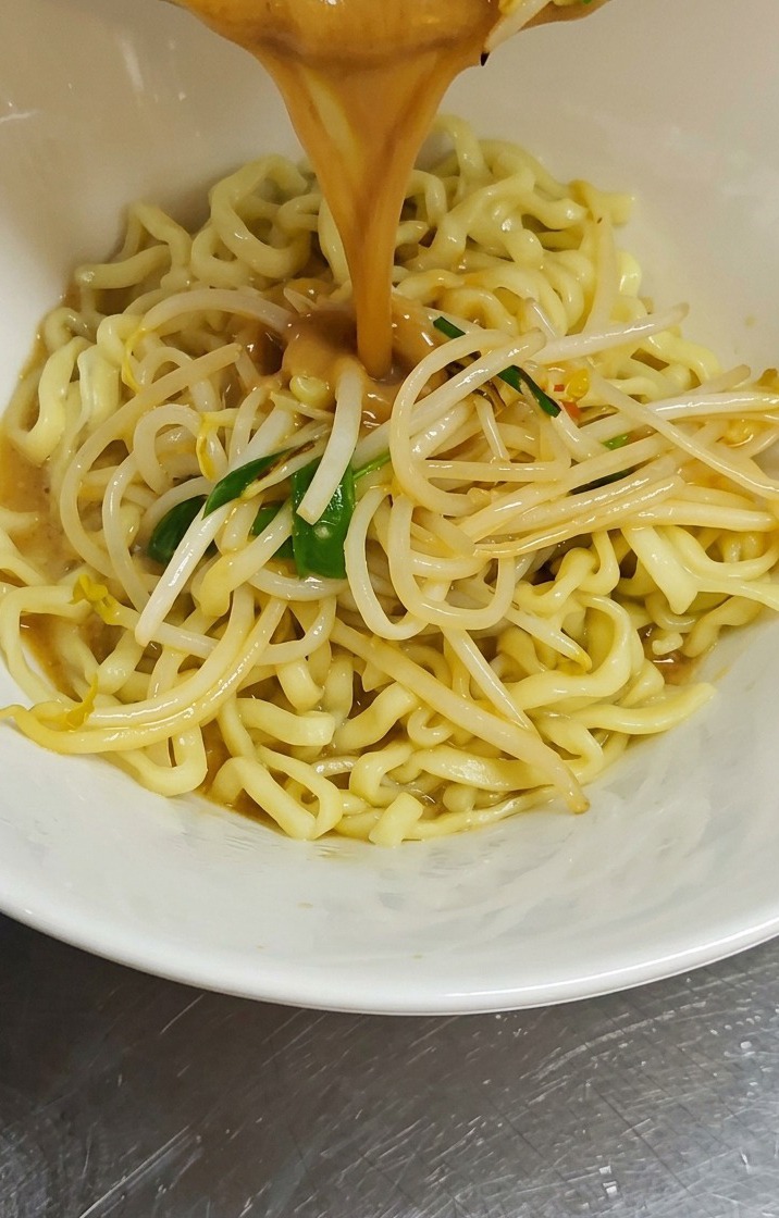 Un flujo de caldo de miso marrón y espeso con brotes de soja salteados se vierte desde una sartén en un tazón blanco lleno de fideos de ramen amarillos y ondulados.