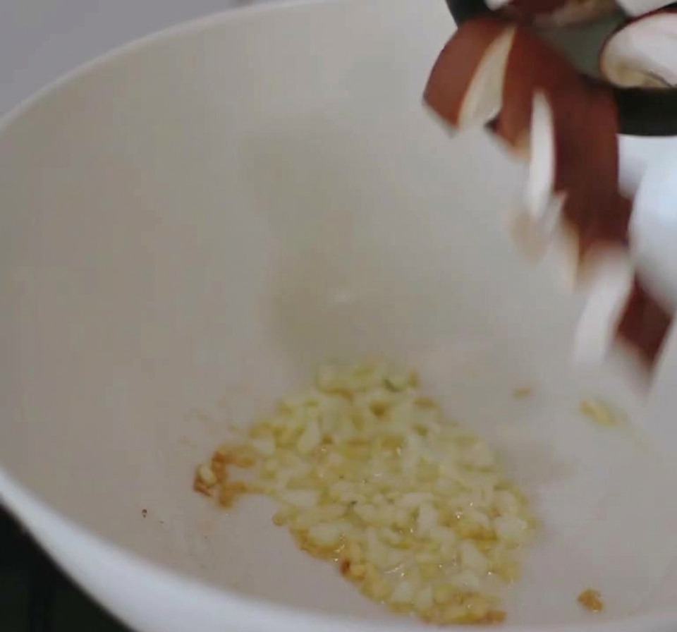 Una sartén blanca en una estufa que contiene ajo picado salteado al que se le añaden champiñones shiitake en rodajas desde arriba.