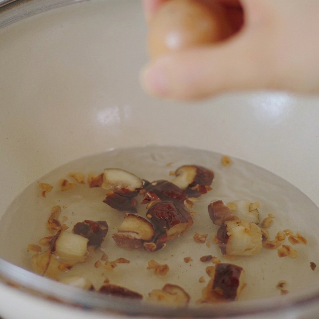 Una mano bajando un huevo de gallina marrón a una olla blanca llena de agua hirviendo y champiñones shiitake en rodajas.