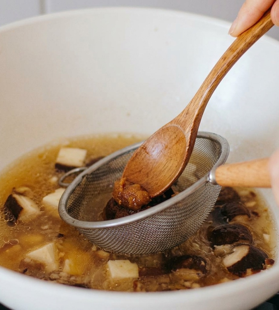 Una cuchara de madera presionando pasta de miso oscura a través de un pequeño colador de metal en un caldo hirviendo a fuego lento con rodajas de champiñones.