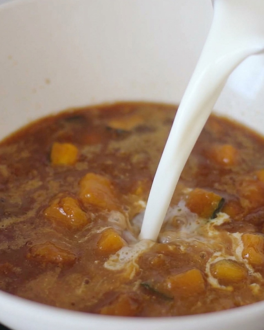 Un chorro de leche blanca siendo vertido en un caldo de miso marrón oscuro que contiene trozos de verduras.
