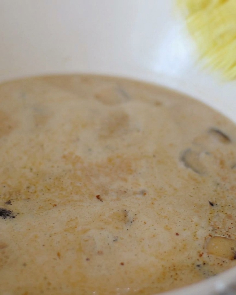 Un bloque de fideos ramen instantáneos amarillos siendo sumergido en una olla de caldo blanco cremoso burbujeante.