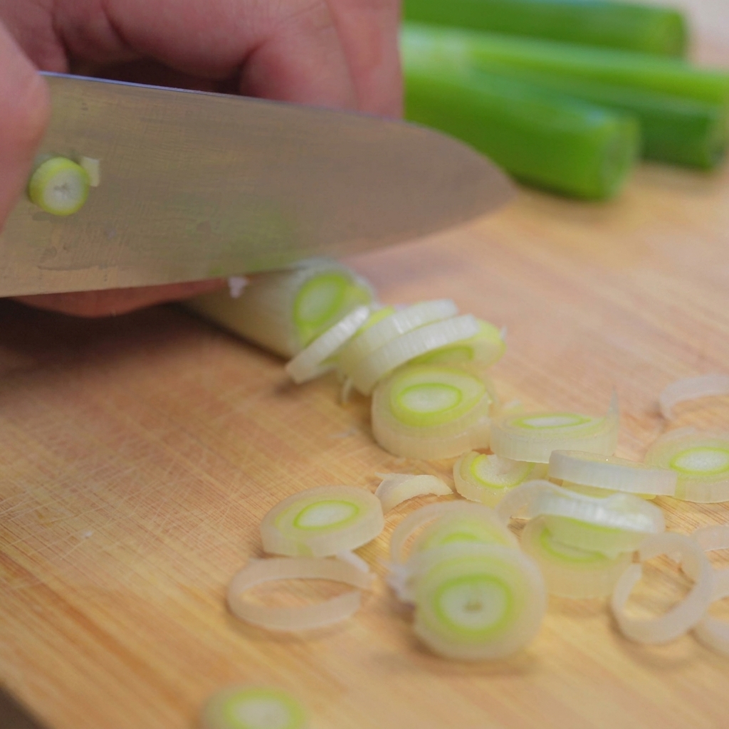 Primer plano de un cuchillo de chef cortando un manojo de cebolletas frescas sobre una tabla de cortar de madera clara.
