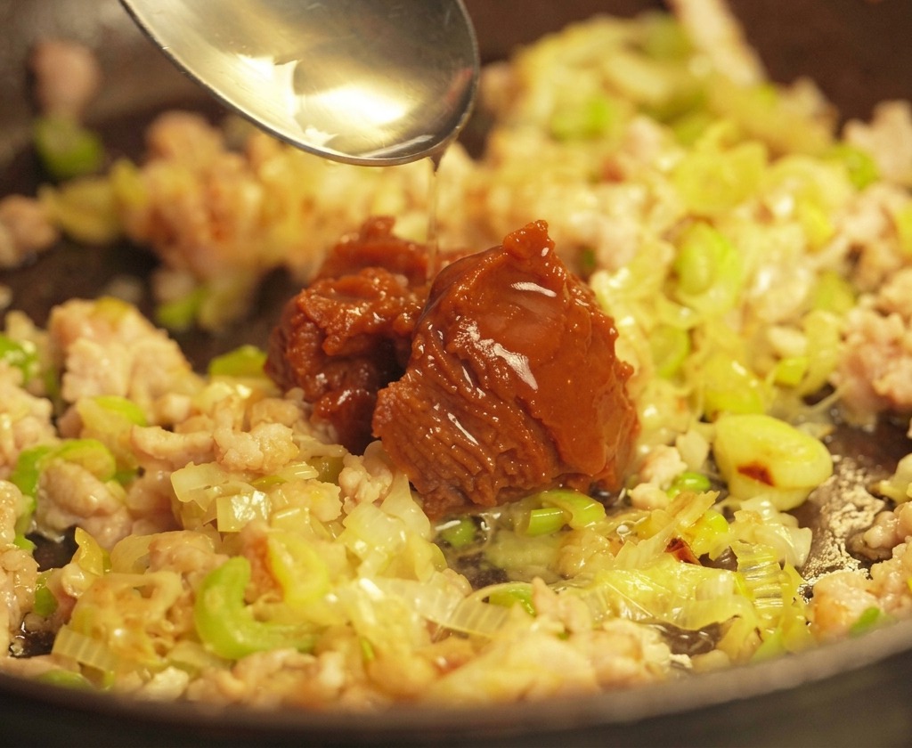 Una cuchara plateada vertiendo una gran cantidad de pasta de miso marrón oscuro sobre una base de carne de cerdo y cebolletas salteadas en una sartén.