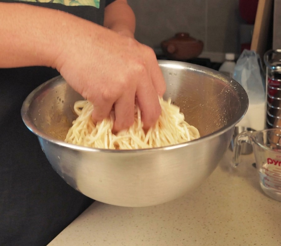 Una persona mezclando a mano una tanda de fideos blancos cocidos en un bol grande de acero inoxidable para cubrirlos con aceite.