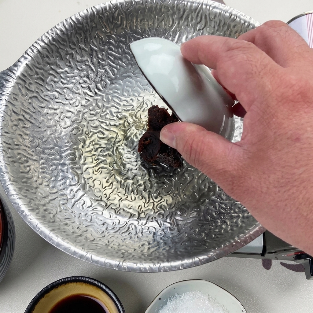 Una mano utilizando un pequeño cuenco blanco para echar una cucharada de pasta de miso rojo oscuro en el líquido reducido del fondo de la olla plateada.