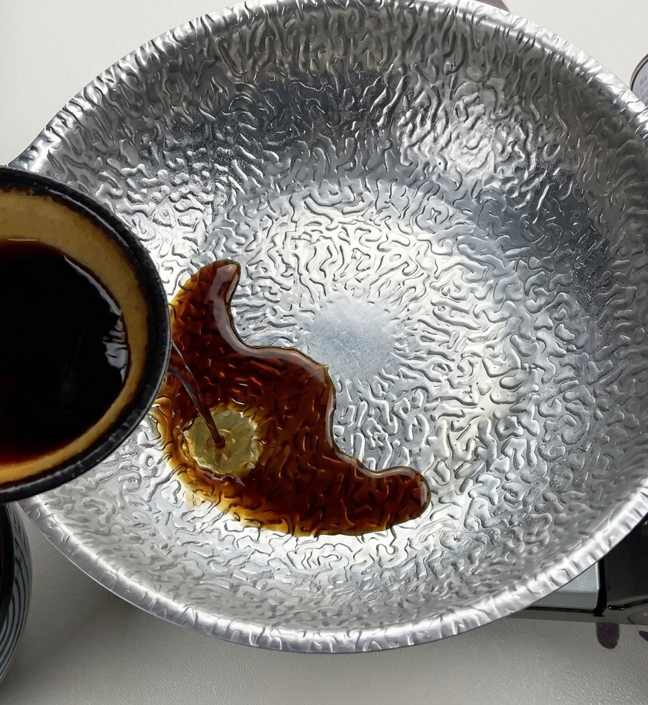 Salsa de soja oscura vertiéndose desde un pequeño cuenco con el interior amarillo a una olla de metal texturizado que contiene miso y azúcar.