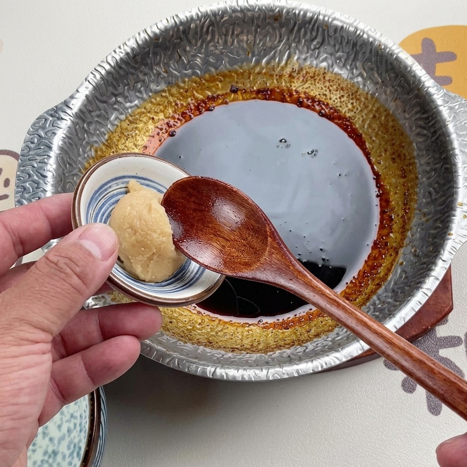 Una cuchara de madera sosteniendo una bola de pasta de miso blanco claro sobre una salsa oscura a fuego lento en una olla de metal.