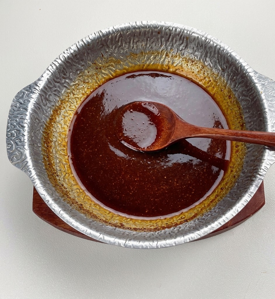 La salsa de miso oscuro terminada cociendo a fuego lento en una olla de metal, con un aspecto espeso y brillante y una cuchara de madera apoyada en el centro.