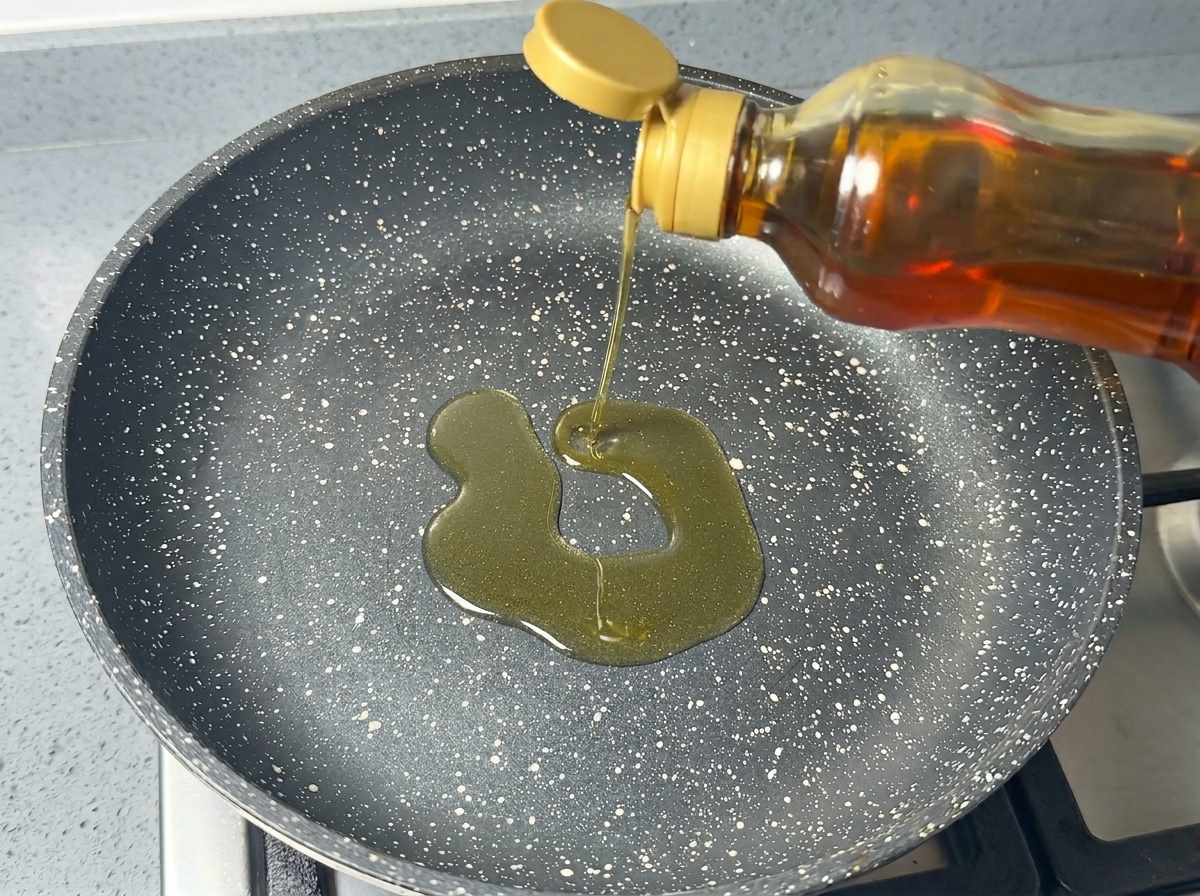 Aceite de cocina vertiéndose desde una botella en una sartén antiadherente oscura y moteada.