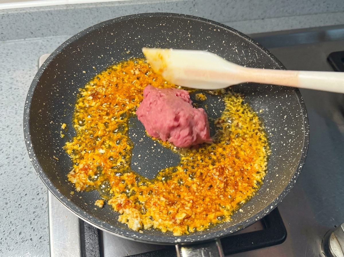 Una porción de carne picada de cerdo cruda y rosada colocándose con una espátula de silicona blanca en una sartén con aromáticos chisporroteantes.
