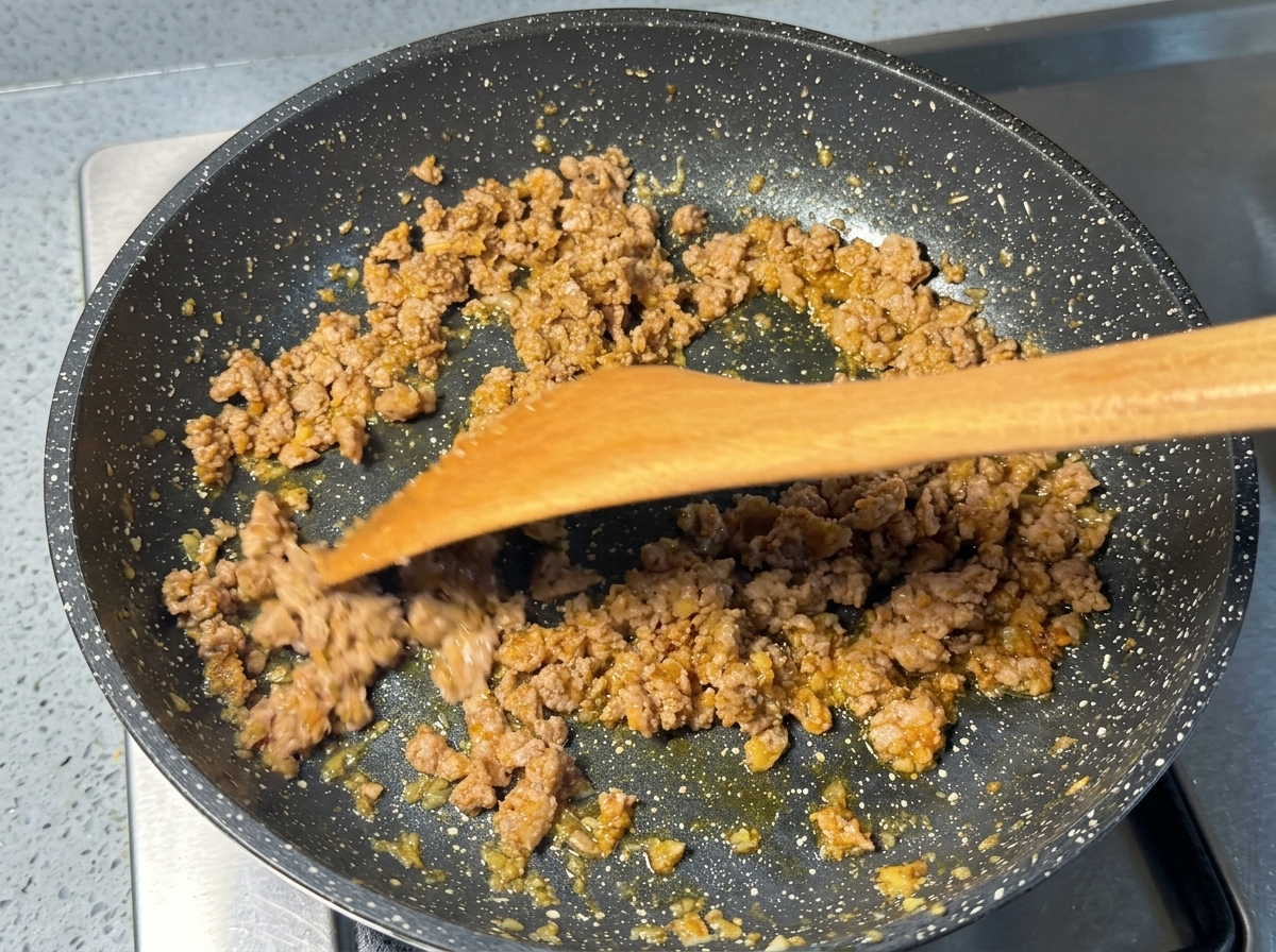 Carne picada de cerdo dorada salteándose en una sartén moteada con una espátula de madera.