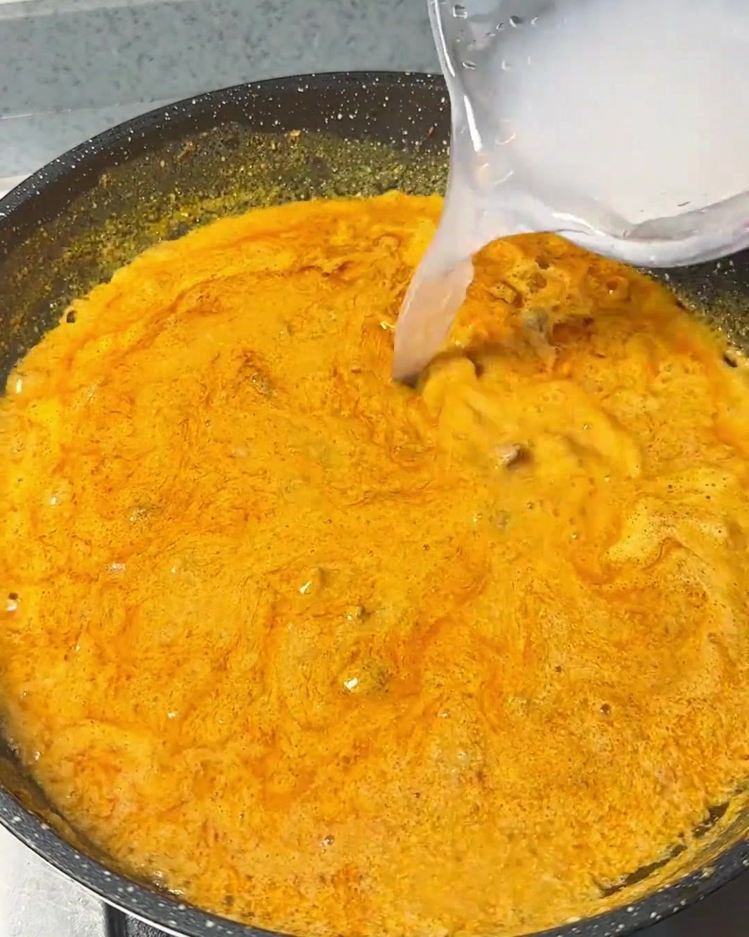 Agua clara vertiéndose desde un recipiente en una mezcla de curry naranja e intensa que burbujea en una sartén oscura.