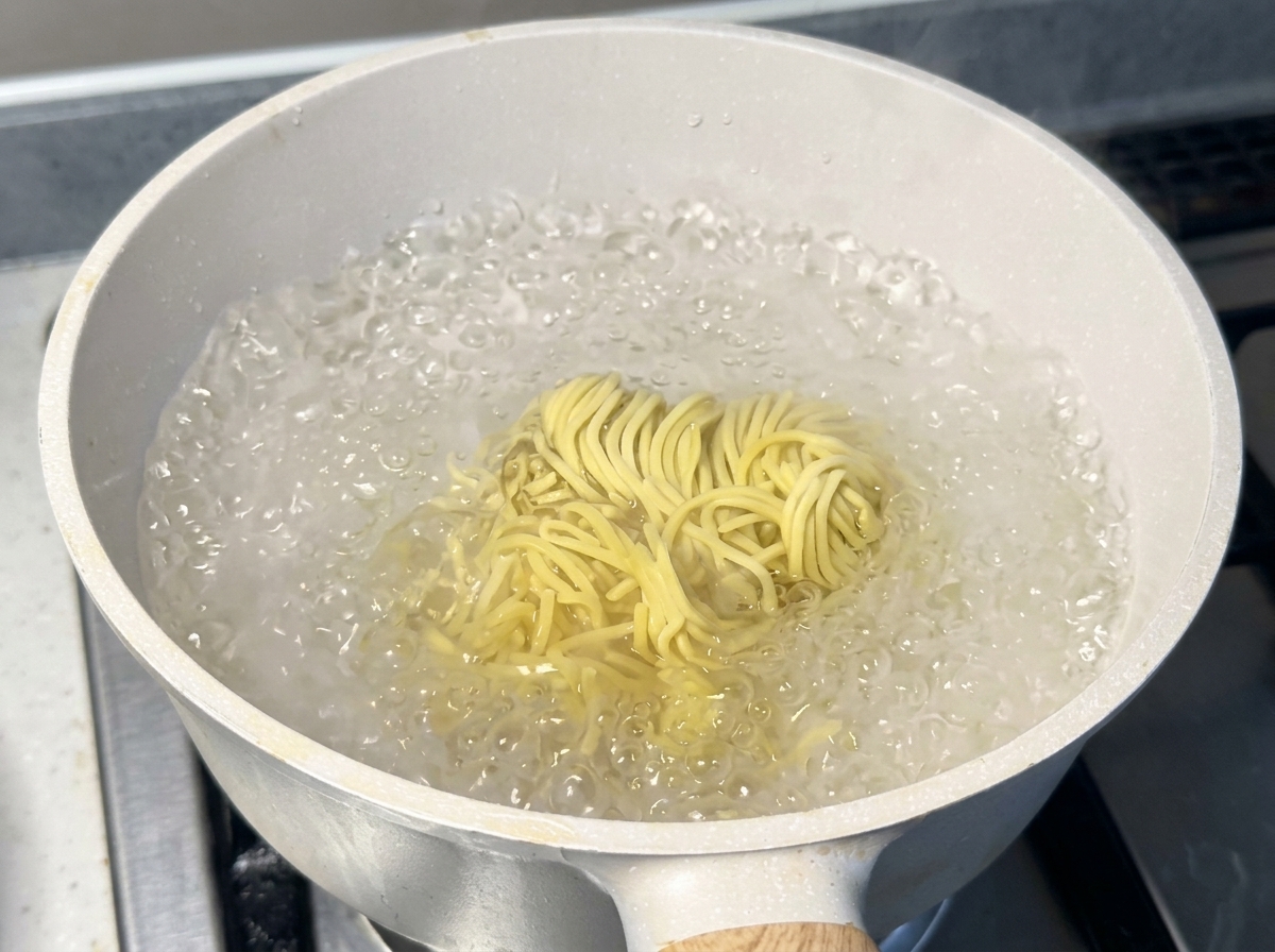 Un nido de fideos ramen amarillos frescos hirviendo vigorosamente en una olla blanca llena de agua caliente con burbujas.