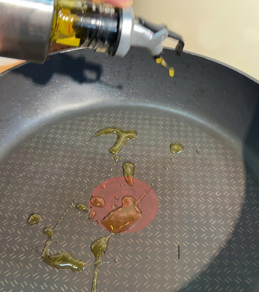 Una mano vertiendo aceite de cocina transparente desde un dispensador de acero inoxidable en una sartén antiadherente negra.