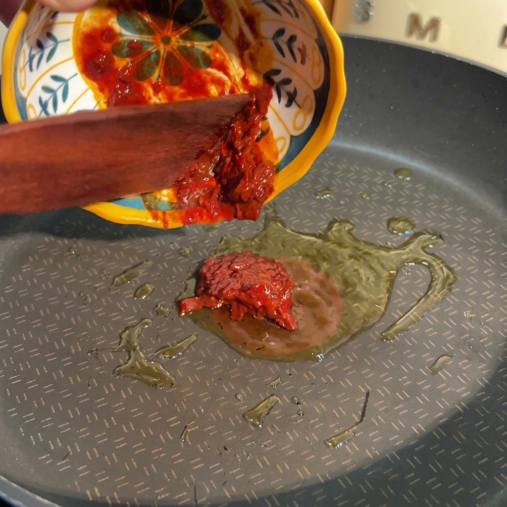 Una espátula de madera empujando pasta roja espesa de frijol desde un tazón decorativo amarillo y azul hacia una sartén con aceite caliente.