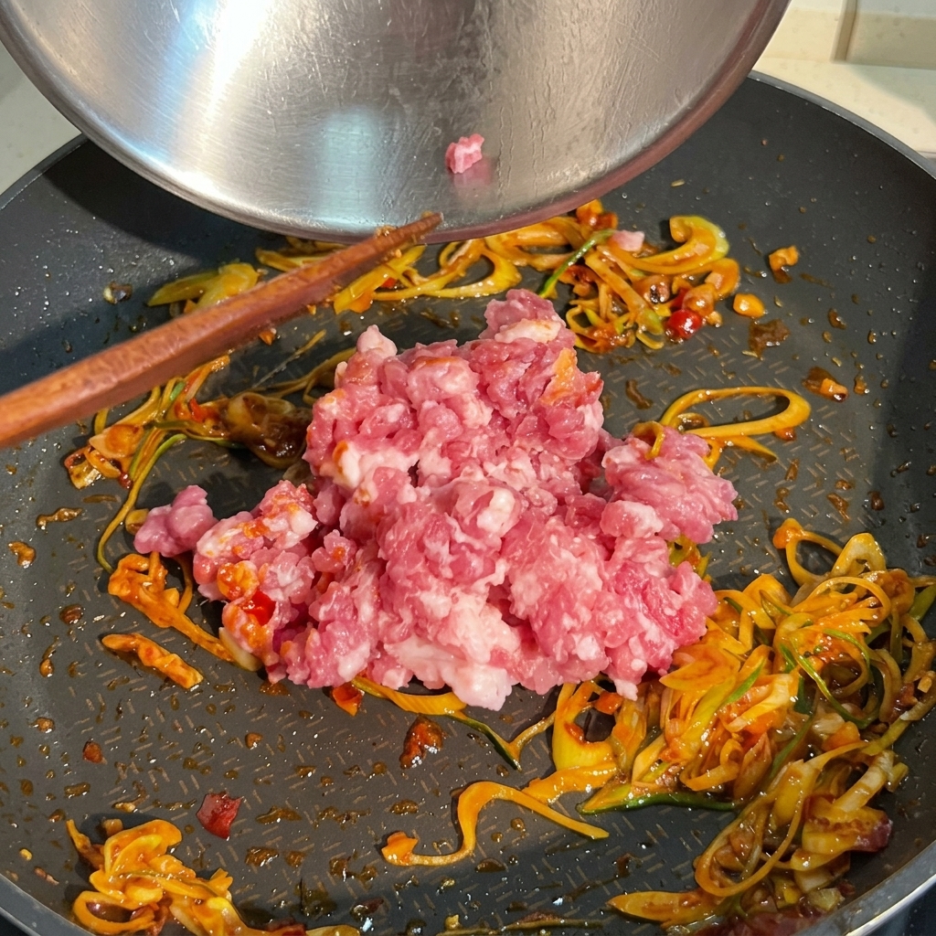 Un montón de carne de cerdo picada cruda y rosada siendo añadida a una sartén que ya contiene cebollines salteados y salsa de chile rojo.