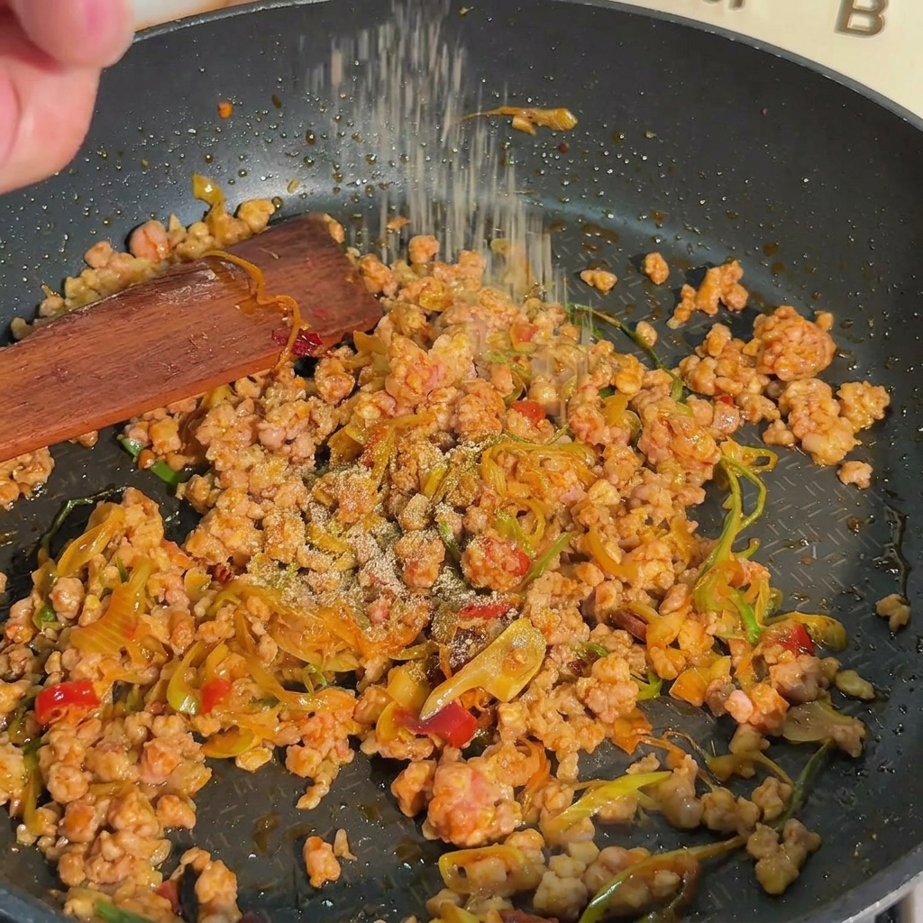 Un primer plano de pimienta blanca espolvoreada sobre carne de cerdo picada dorada y cebollines en una sartén negra.