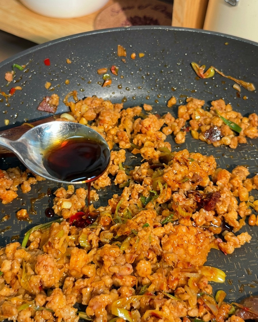 Un líquido oscuro y sabroso vertiéndose desde una cuchara plateada sobre una mezcla de carne picada dorada y cebollines en una sartén.