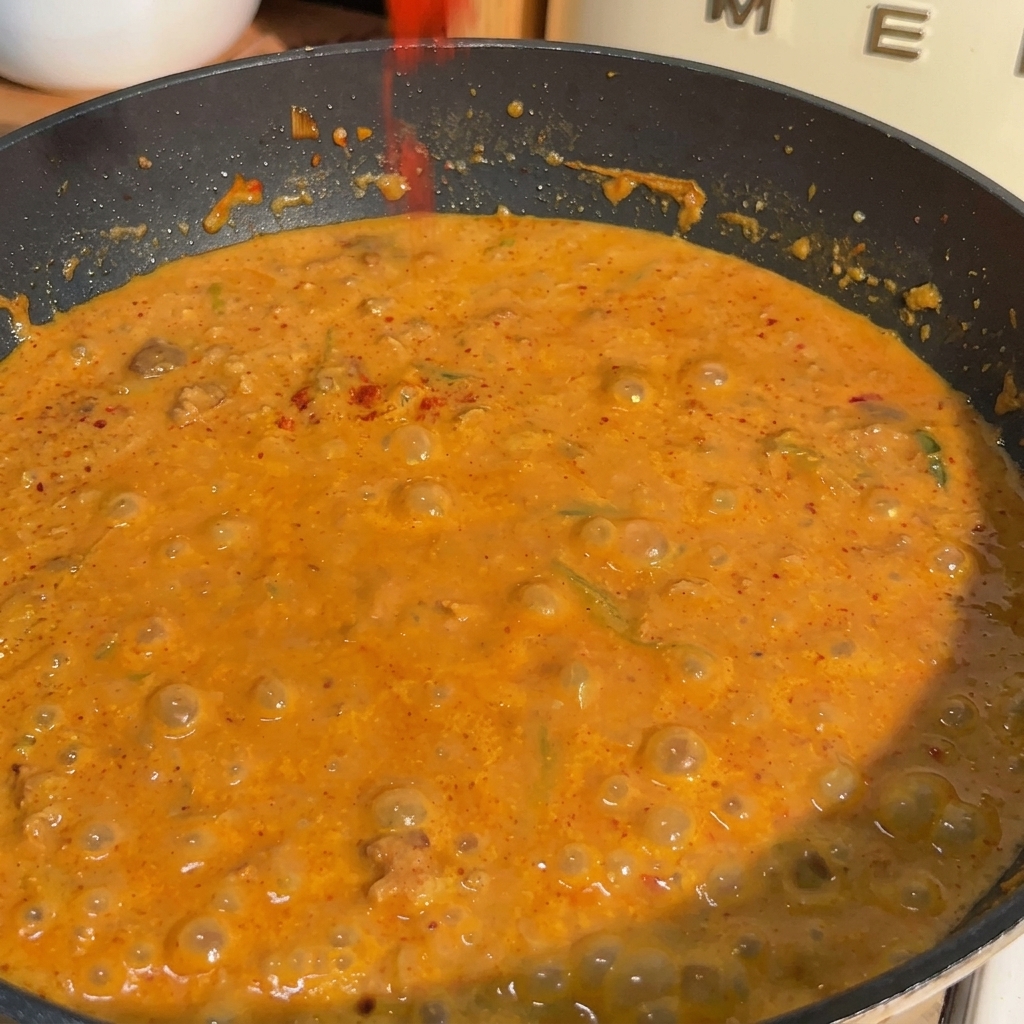 Una sartén llena de salsa de leche de soya y miso de color naranja burbujeando mientras se espolvorea chile rojo en polvo sobre la superficie.