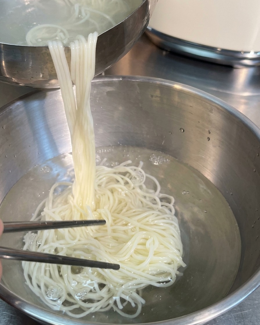 Palillos plateados levantando una gran porción de fideos blancos cocidos desde un tazón de metal con agua hacia otro tazón.