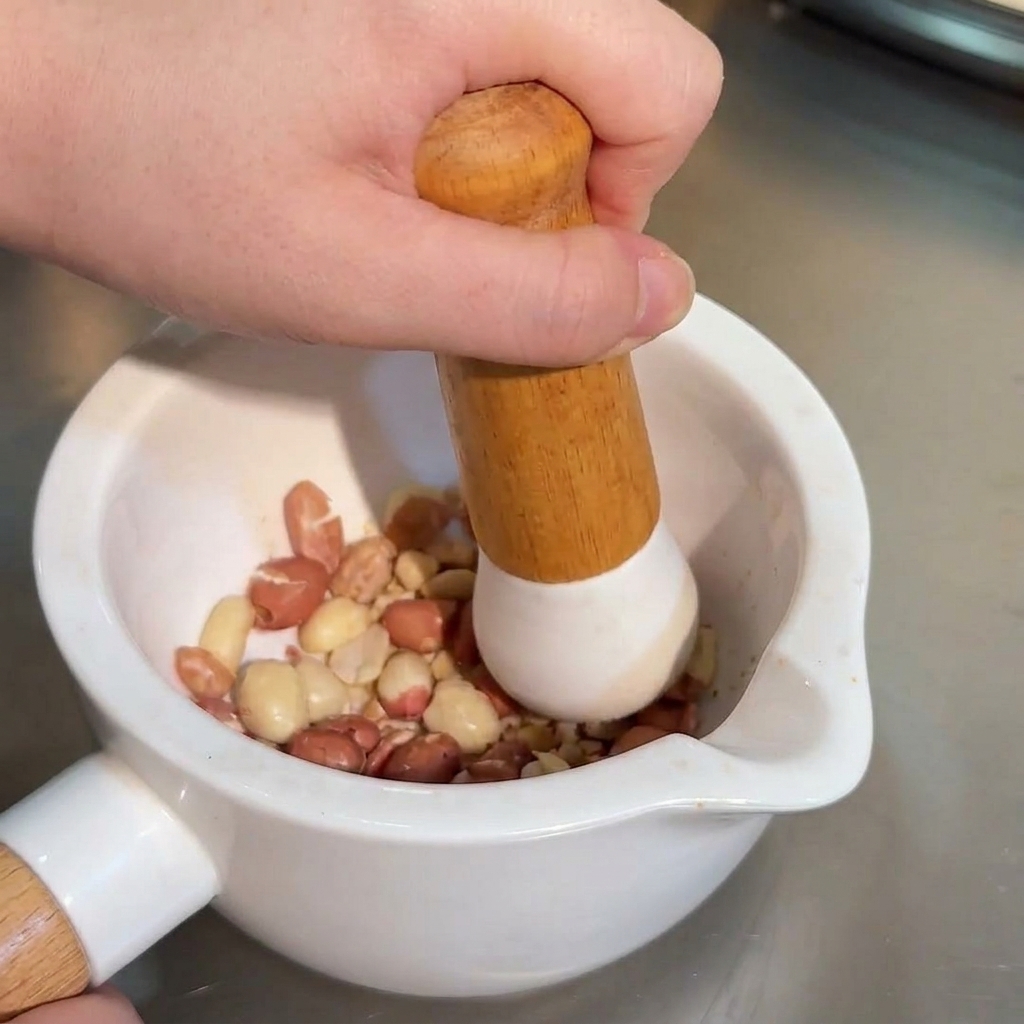 Una mano usando un mazo de madera para triturar cacahuates tostados enteros dentro de un mortero de cerámica blanca.