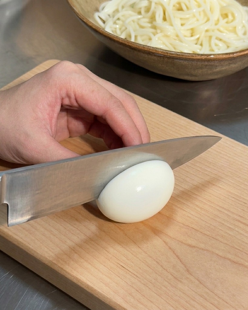 Una mano sosteniendo un cuchillo para cortar un huevo duro tierno sobre una tabla de cortar de madera con un tazón de fideos al fondo.