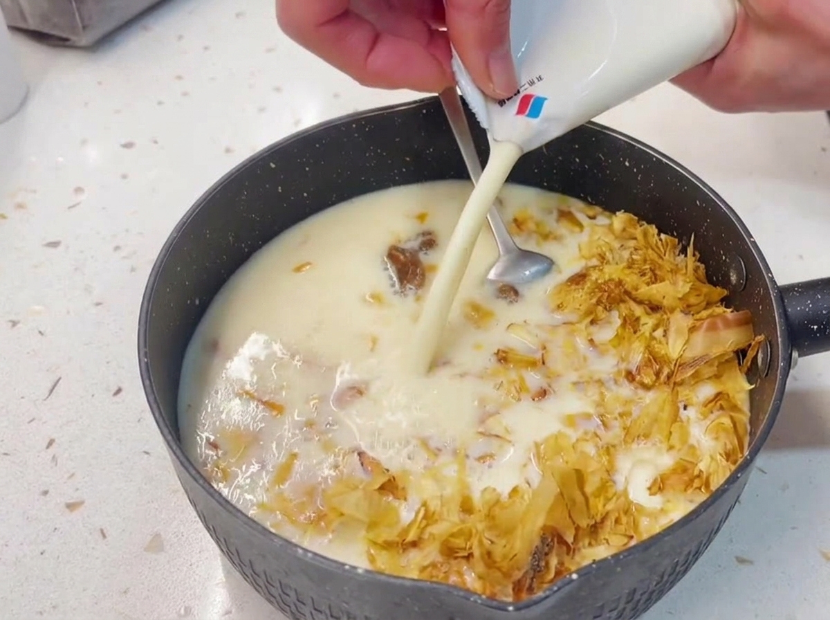 Manos vertiendo leche de soja blanca y cremosa desde un brik hacia un cazo oscuro que contiene una mezcla de atún y copos de bonito seco.