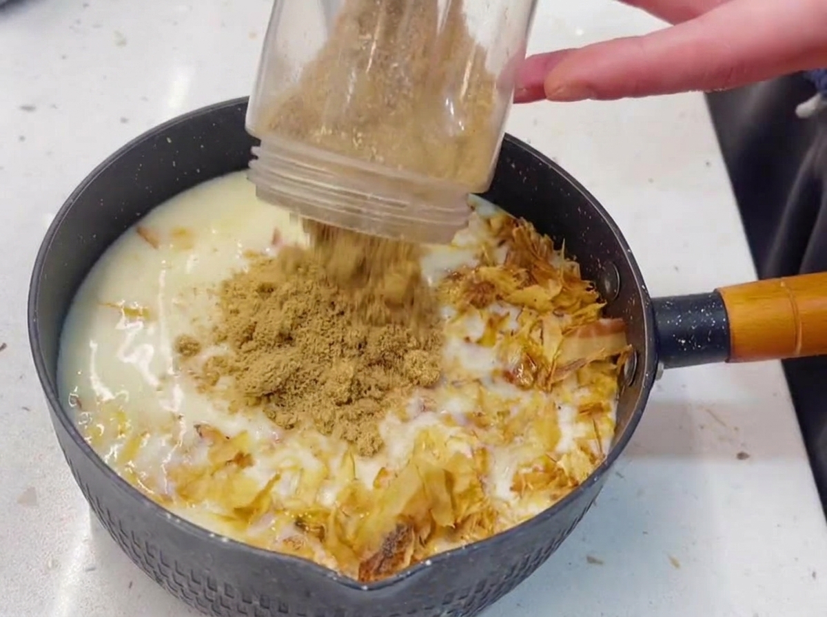 Un vaso de batidora de plástico vertiendo semillas de sésamo tostadas y finamente molidas en una olla con un líquido blanco lechoso, atún y copos de bonito.