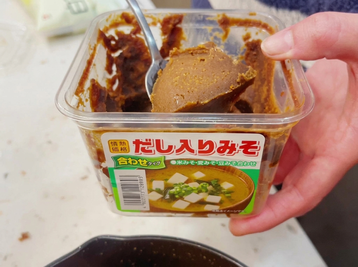 Una mano sacando una cucharada generosa de pasta de miso marrón y espesa de un envase de plástico transparente con una cuchara de metal.