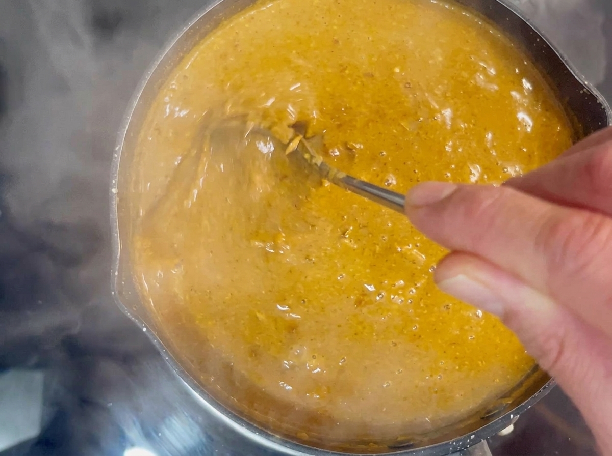 Una mano usando una cuchara de metal para remover un caldo de inmersión espeso y burbujeante de color marrón anaranjado en una olla pequeña.