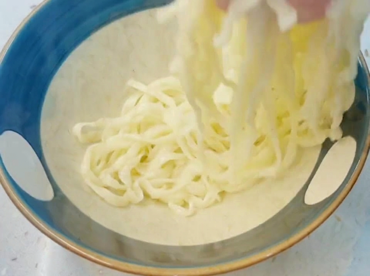 Una mano levantando y moviendo fideos de ramen blancos, gruesos y cocidos dentro de un bol azul lleno de agua fría.