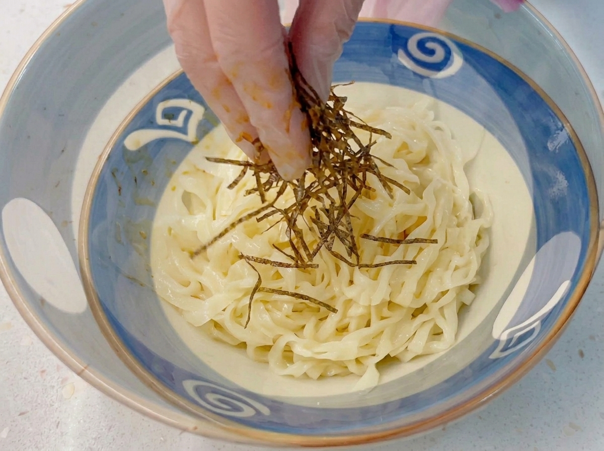Dedos con guantes dejando caer alga nori en tiras sobre un bol de fideos de ramen gruesos y fríos.