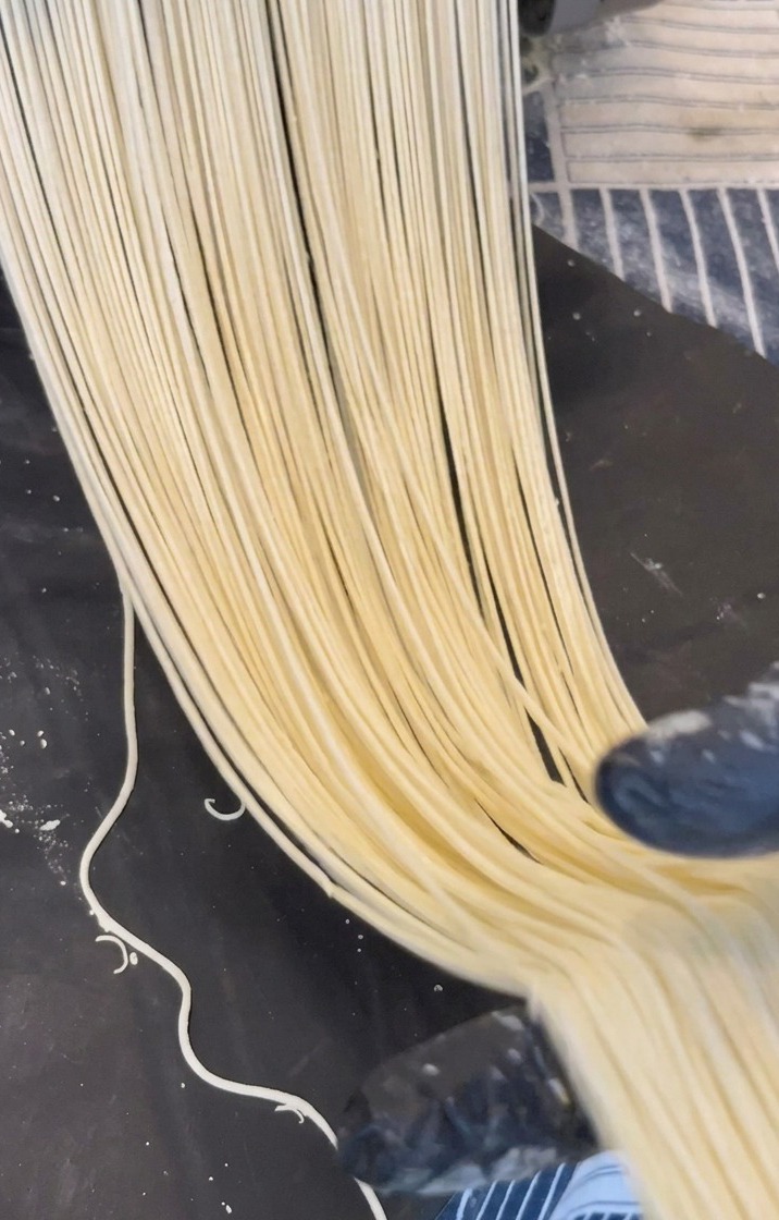 Hebras largas de fideos ramen de color amarillo pálido saliendo de una máquina metálica sobre una superficie oscura.