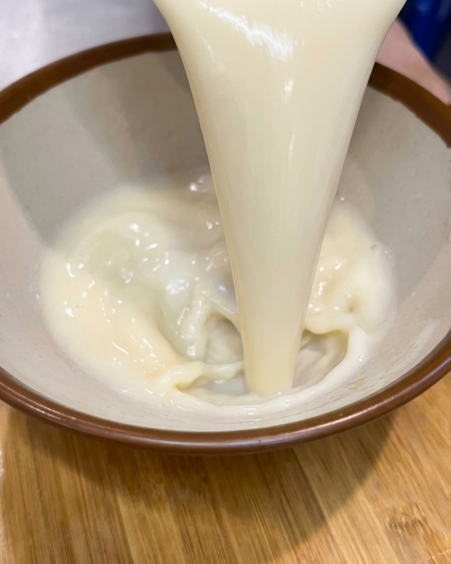 Un chorro espeso de base de sopa cremosa y pálida vertiéndose en un cuenco de cerámica con borde marrón.