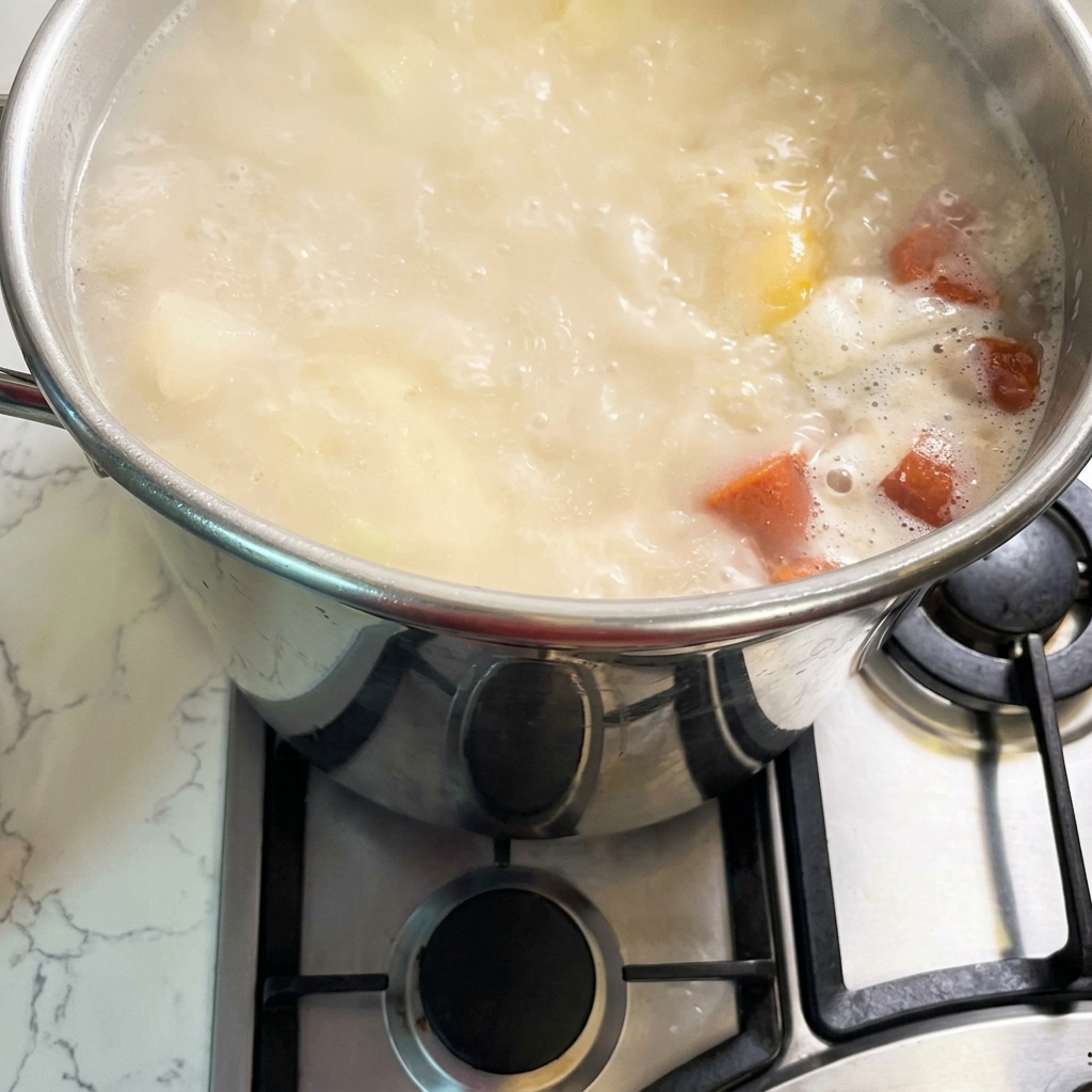 Una olla grande de acero inoxidable sobre un fogón de gas llena de un caldo burbujeante de color blanco cremoso y naranja.