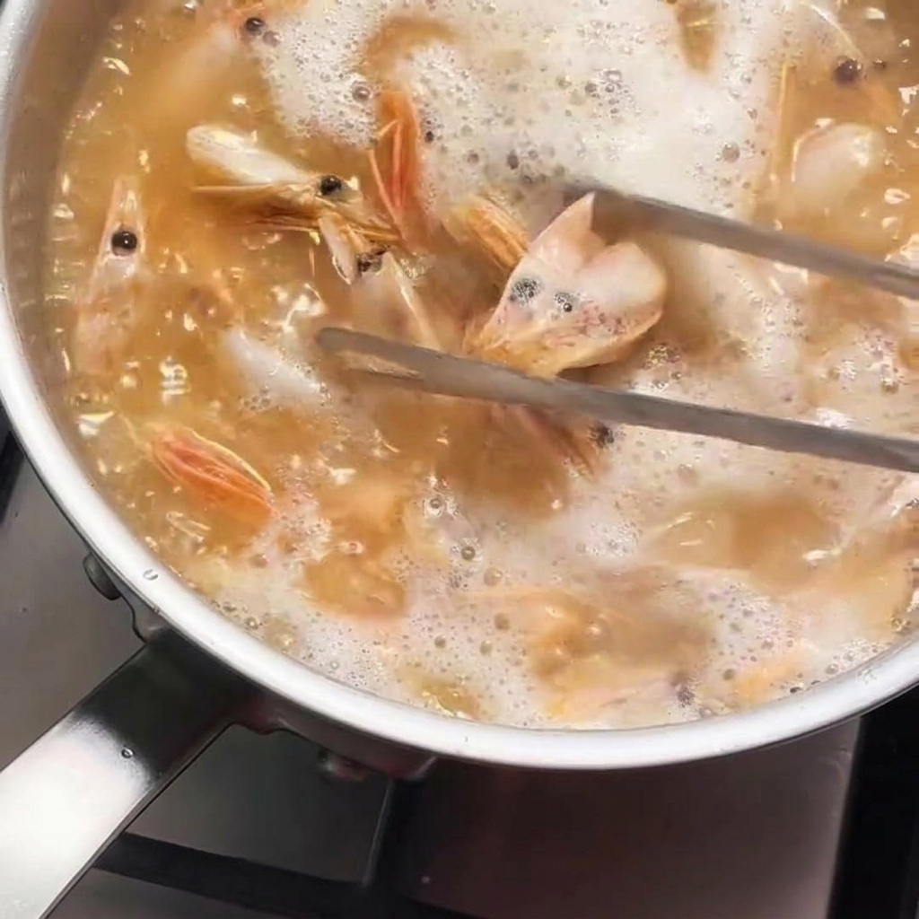 Cabezas de camarón removidas con pinzas metálicas en una cazuela con aceite caliente y burbujeante que va adquiriendo un color naranja intenso.