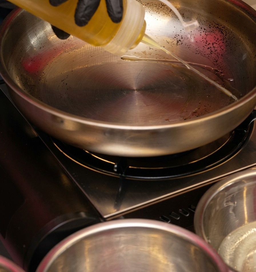 Aceite de cocina transparente vertiéndose desde una botella de plástico hacia una sartén de acero inoxidable reluciente sobre un quemador portátil.