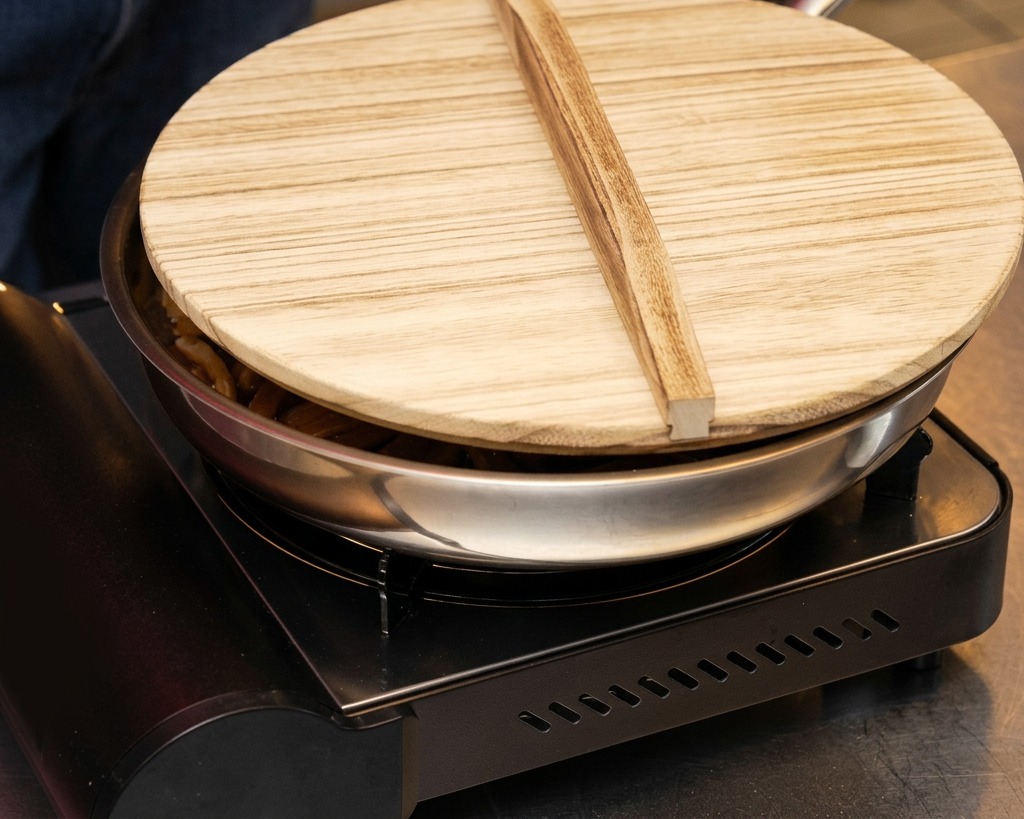 Una sartén de acero inoxidable con brotes de bambú sobre una estufa de gas portátil, cubierta con una tapa de madera tradicional.