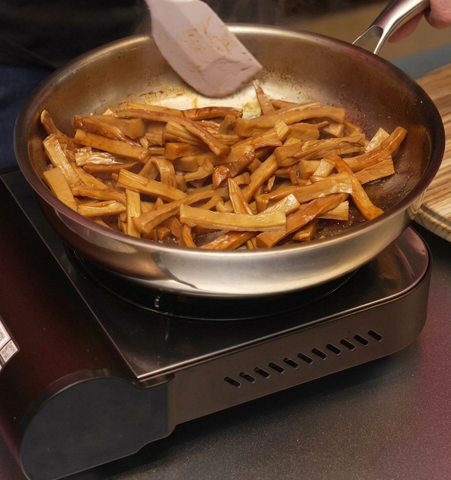 Una espátula blanca removiendo brotes de bambú marrones y glaseados en una sartén para asegurar que queden cubiertos por el aromático aceite de sésamo.