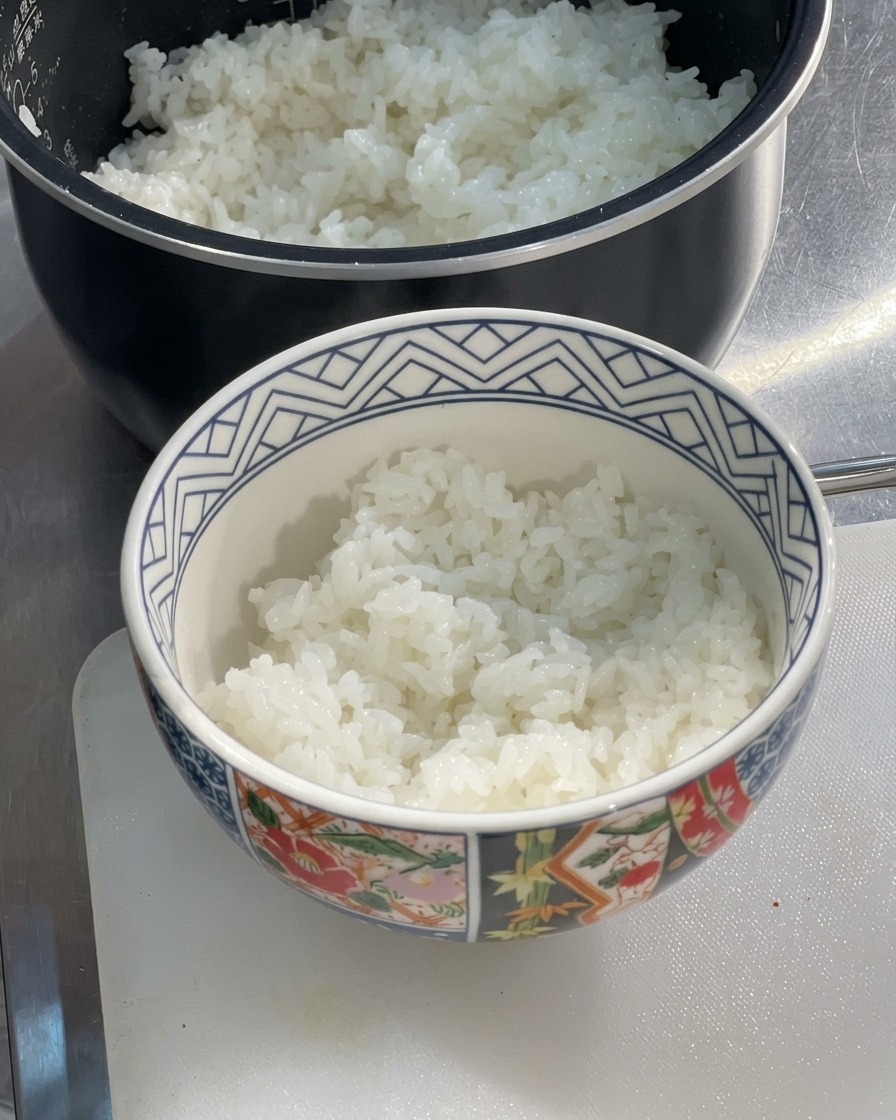 Un tazón de cerámica con motivos lleno hasta la mitad con arroz blanco recién hecho al vapor y esponjoso, asentado sobre una tabla de cortar junto a una arrocera.