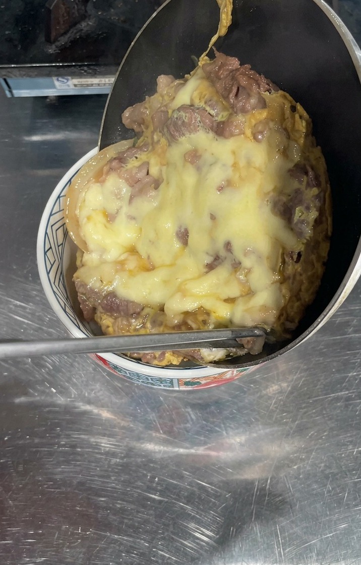 Una sartén inclinada sobre un tazón de arroz blanco, con una gruesa capa de ternera cocinada y queso amarillo fundido deslizándose para cubrir la superficie.
