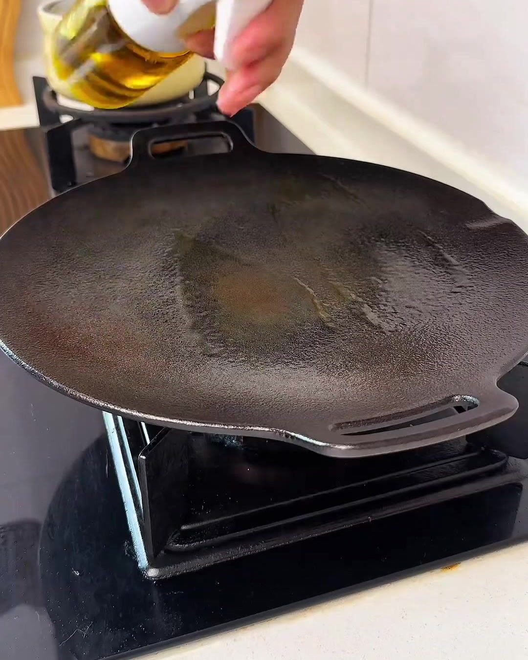 Una mano rociando una fina bruma de aceite de cocina dorado sobre una plancha circular negra y limpia en la estufa.