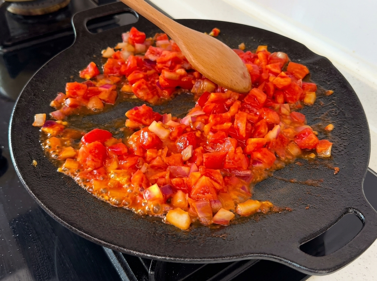 Tomates machacados y troceados mezclándose con cebollas en una sartén caliente, mostrando sus jugos burbujeando de forma visible.