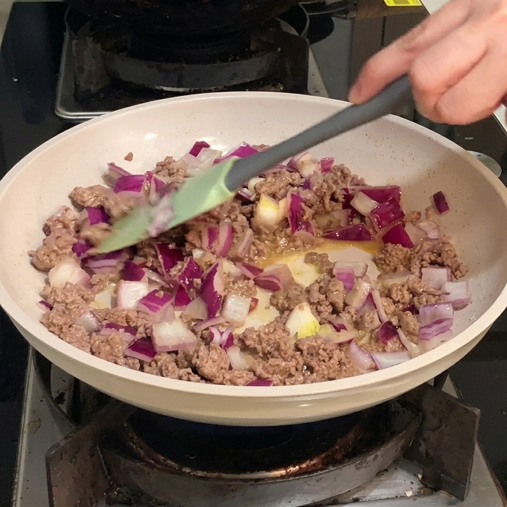 Dados de cebolla morada mezclándose con carne picada dorada en una sartén.