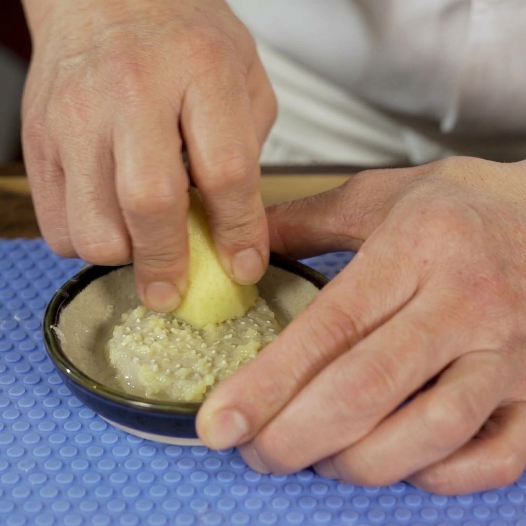 Una persona utilizando un rallador de cerámica para convertir un trozo de raíz de jengibre fresco en una pasta fina en un bol pequeño.